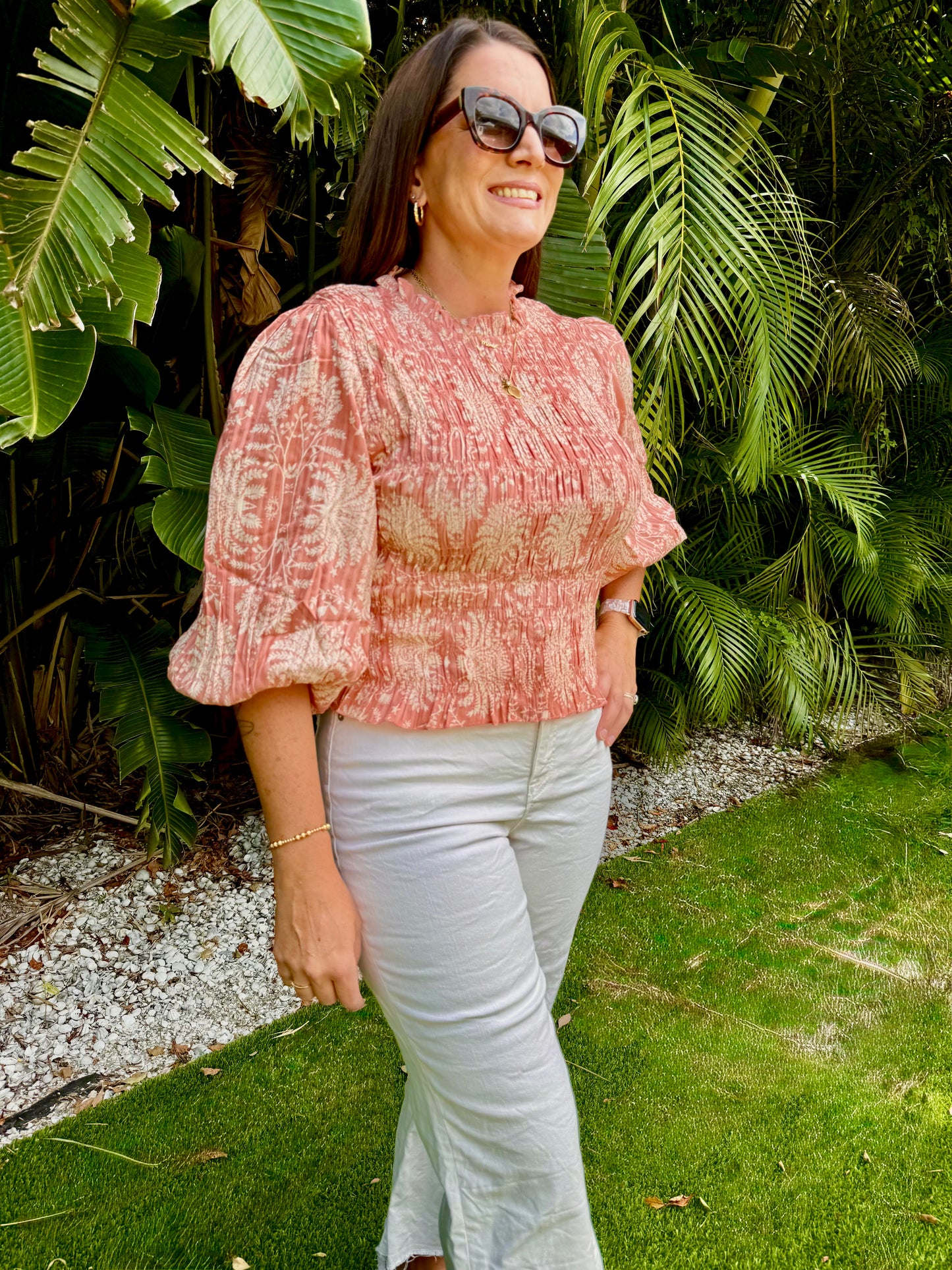 Woman wearing a pink patterned blouse and white jeans standing in front of green foliage.