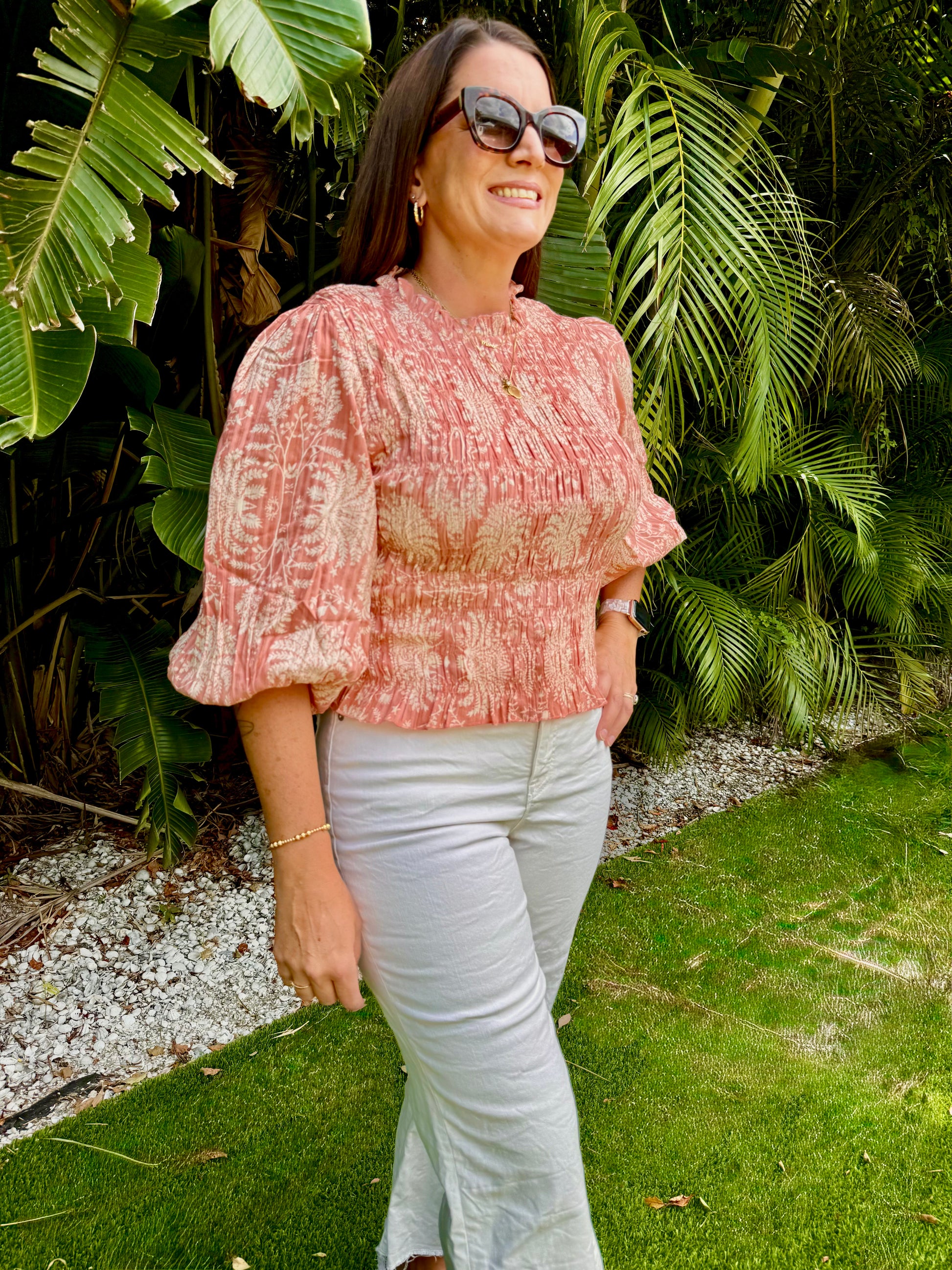 Woman wearing a pink patterned blouse and white jeans standing in front of green foliage.